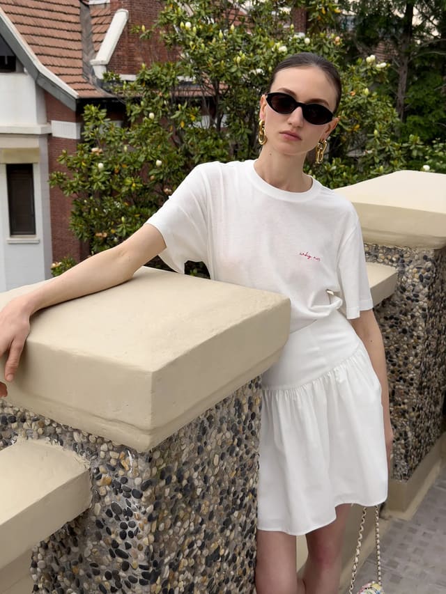 Model wearing white relaxed t-shirt with red 'why not' embroidery, paired with white tiered skirt, outdoors on balcony