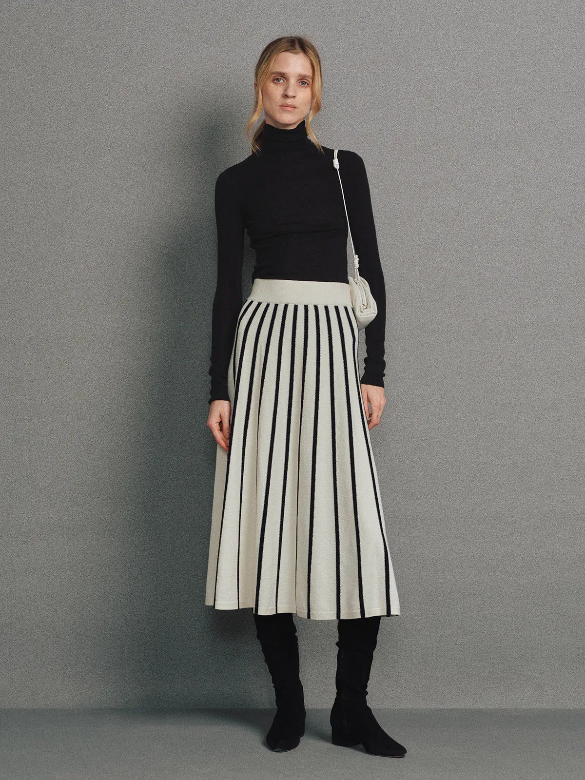 Front view of a model wearing a Black/White vertical stripe A-line knit skirt with white waistband, black turtleneck, and black boots.