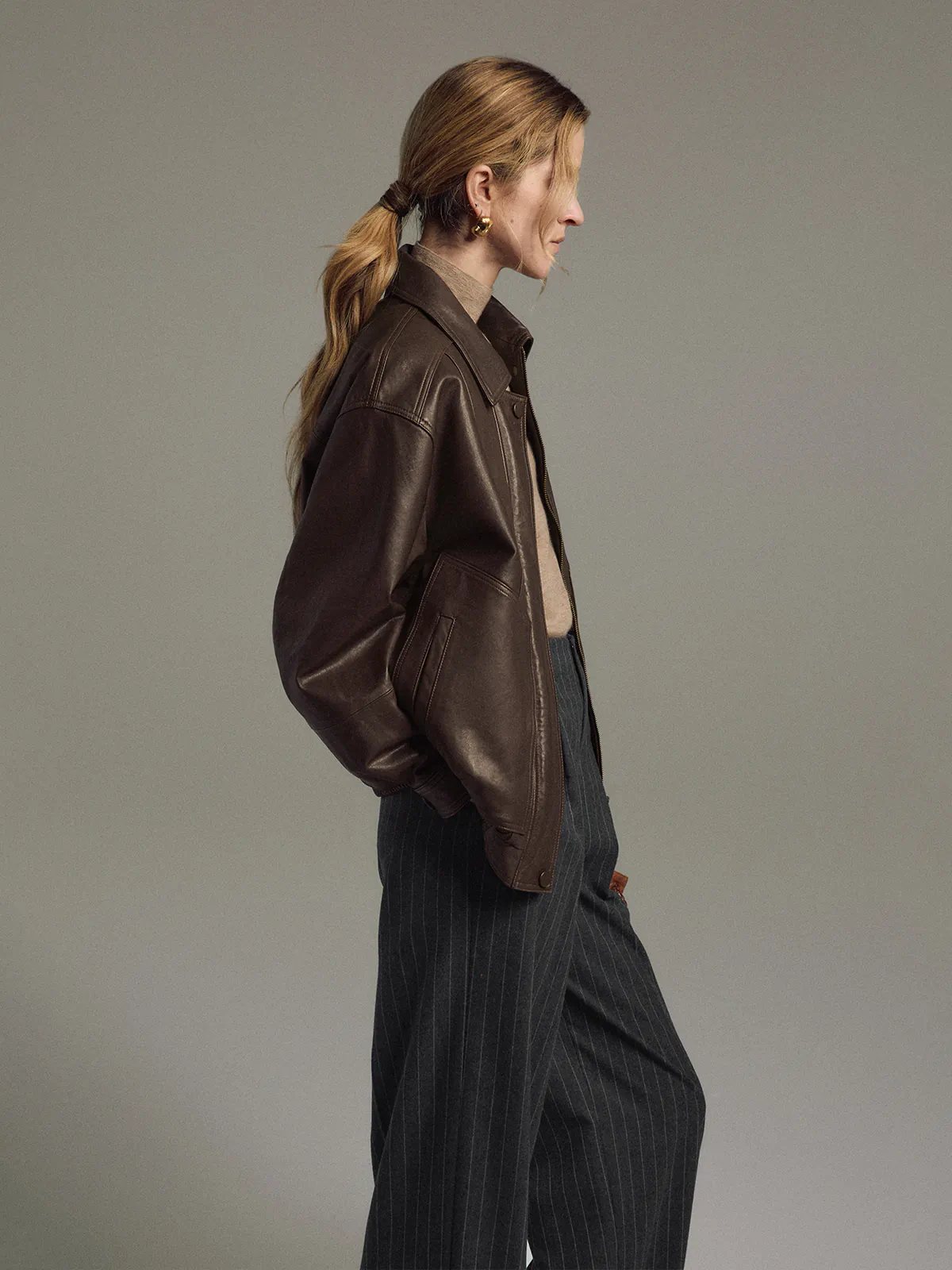Model in a Dark Brown Italian shearling leather jacket, side view, holding a tote bag.