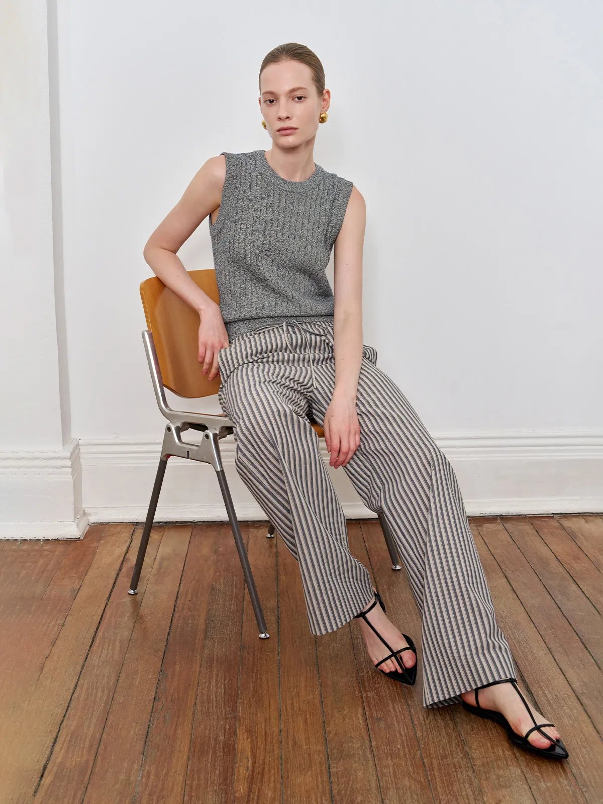Model seated on mid-century chair wearing grey MILLIE knit tank top with striped wide-leg trousers — minimalist fashion editorial