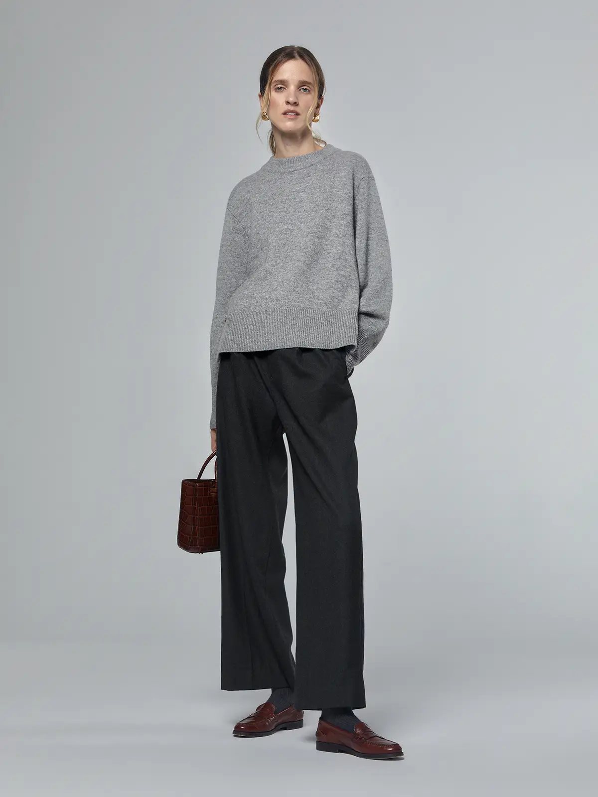 Model wearing a grey merino wool sweater with metal buttons and ribbed hem, styled with dark trousers and loafers.