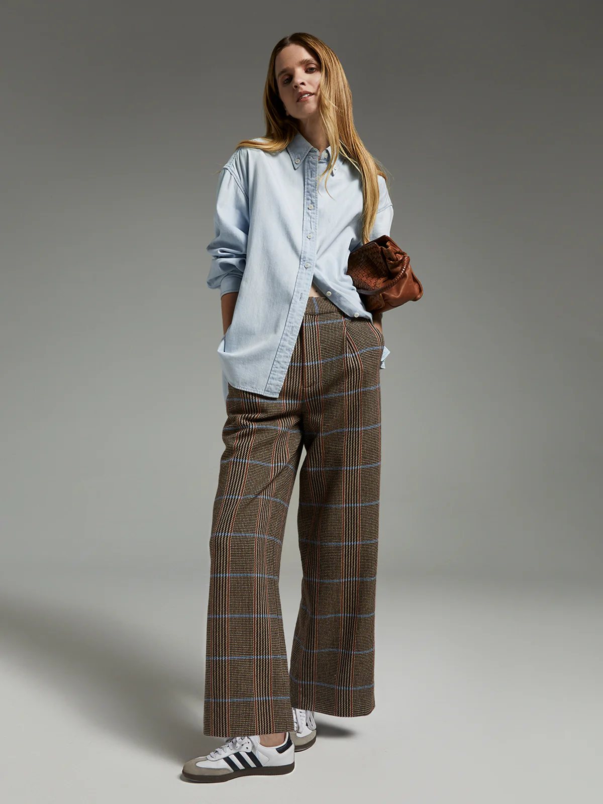 Model wearing brown wool British check trousers with wide legs, pleats, and hands in pockets, paired with a top and sneakers.