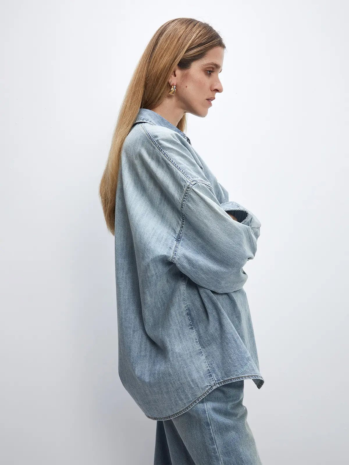 Side profile of a model wearing a light blue oversized denim shirt with dropped shoulders and raw hem, arms crossed.
