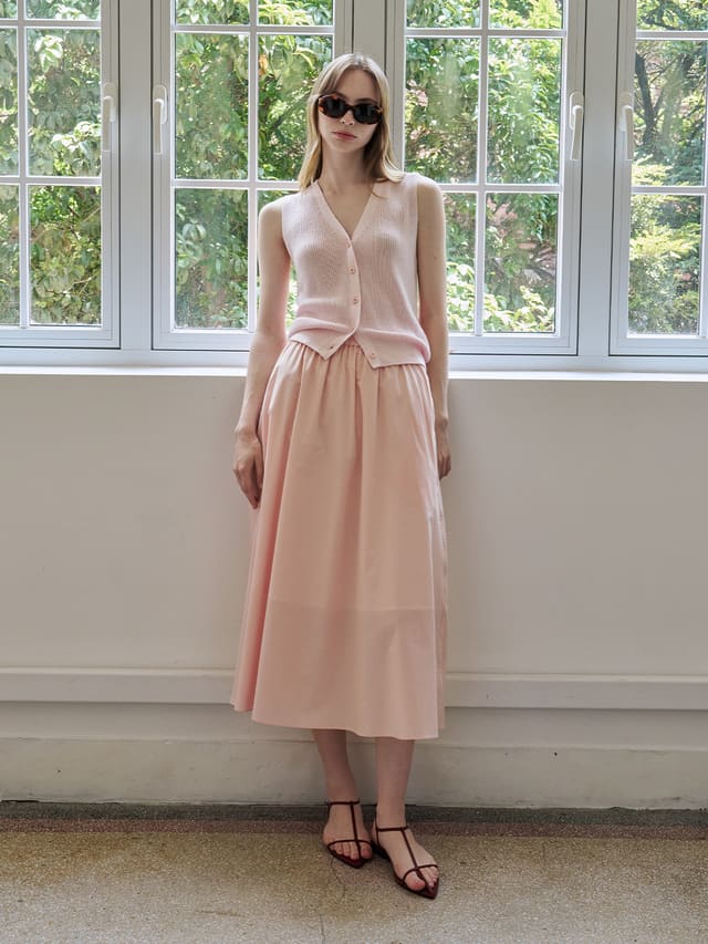 Andreatang blush lyocell cotton A-line maxi skirt worn with pink knit vest and burgundy strappy sandals, front full-body shot by window