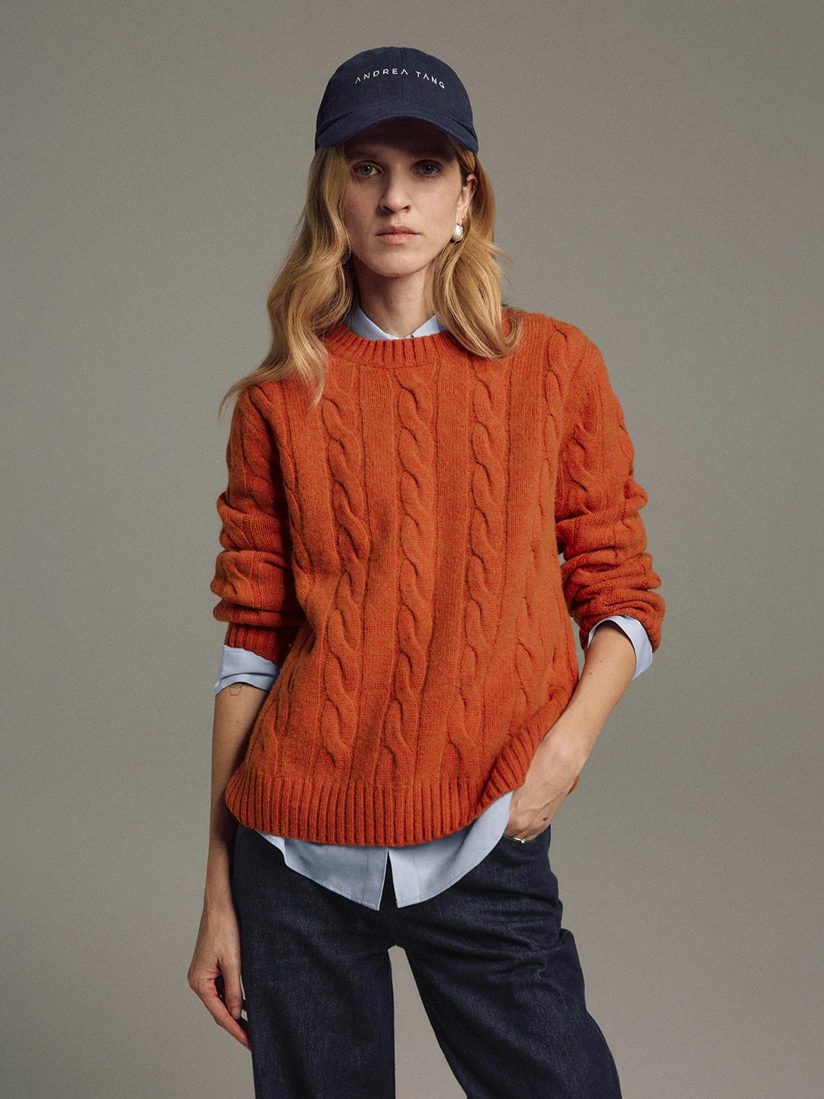 Model wearing an orange cable knit jumper layered over a collared shirt with rolled sleeves, paired with dark jeans and a cap.