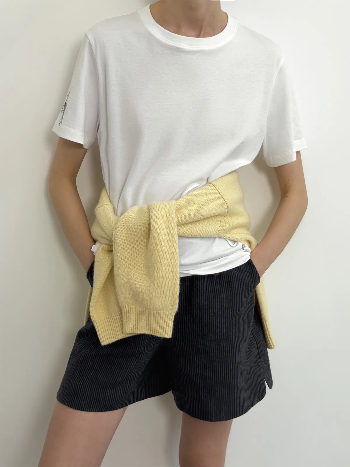 Model wearing NEVAEH relaxed linen shorts with white tee and knotted yellow cardigan, hands in pockets