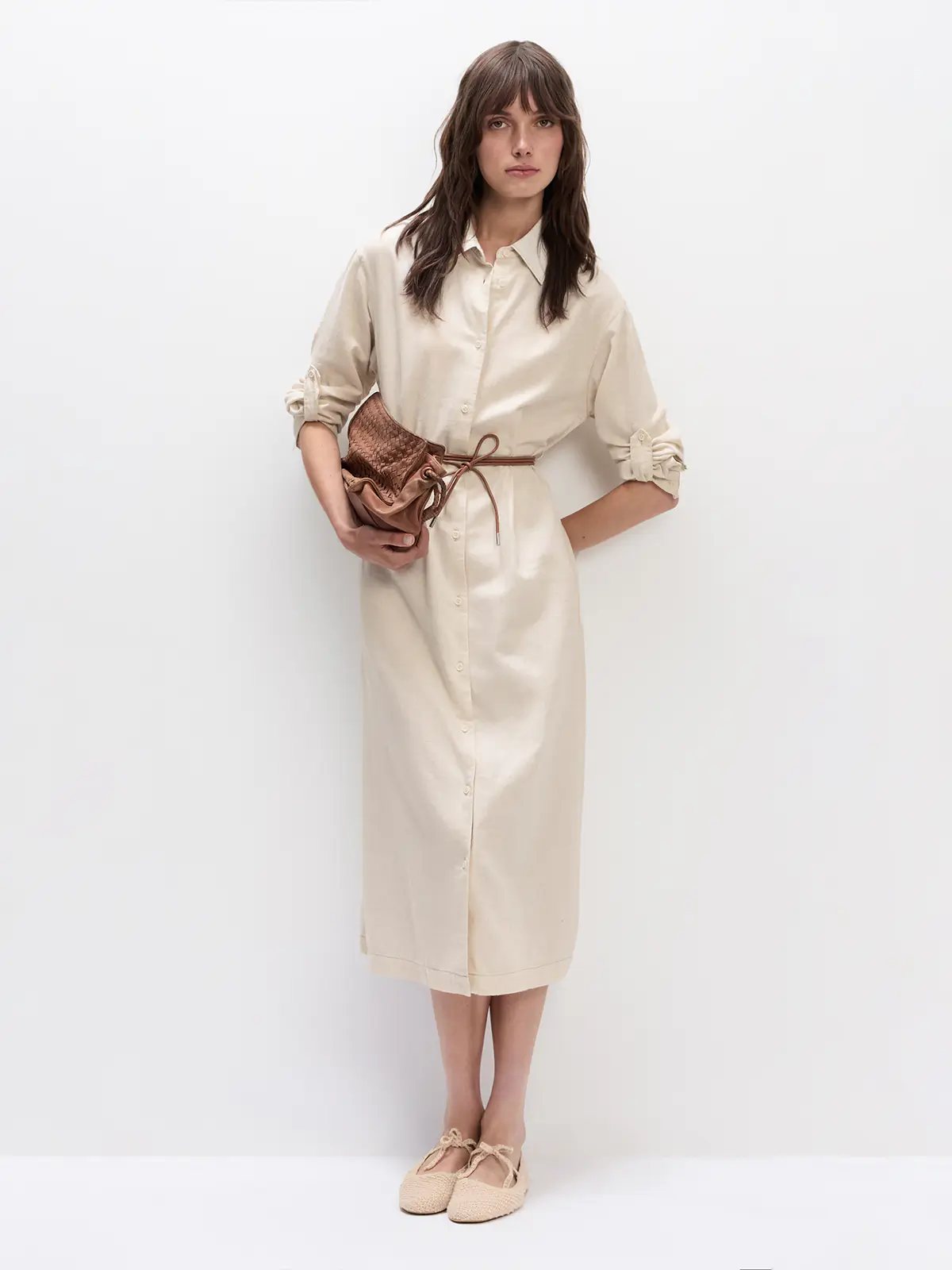 Front view of the Natural JOANNA shirt dress with rolled sleeves, front buttons, and self-fabric tie belt, holding a woven clutch.