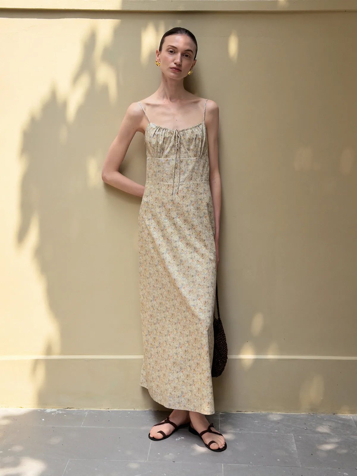 Front full-body shot of model in beige floral PIPPA dress against sun-dappled beige wall, holding beaded tote