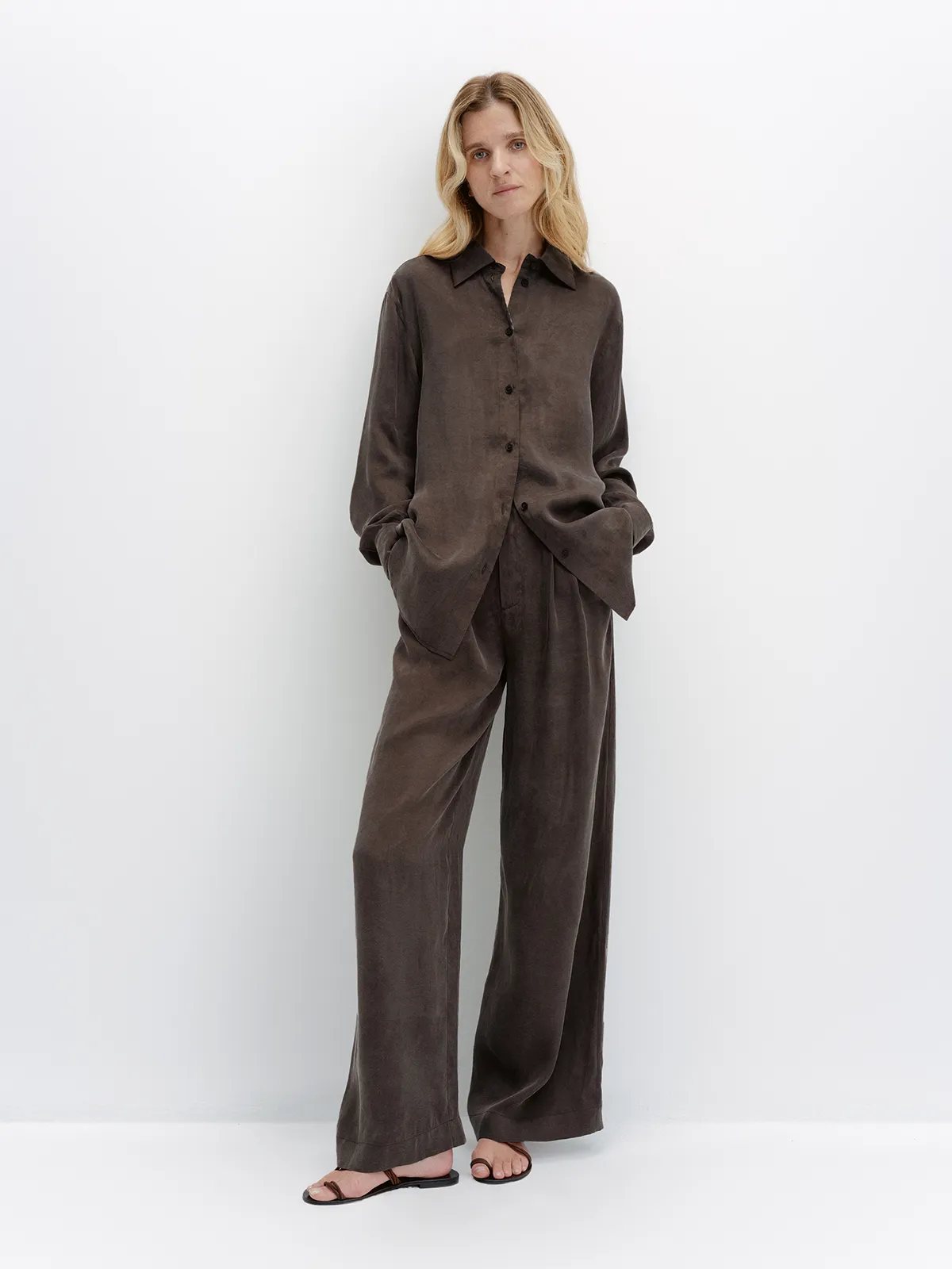 Blonde model in taupe NOBU Trousers and matching shirt, hands in pockets, white backdrop