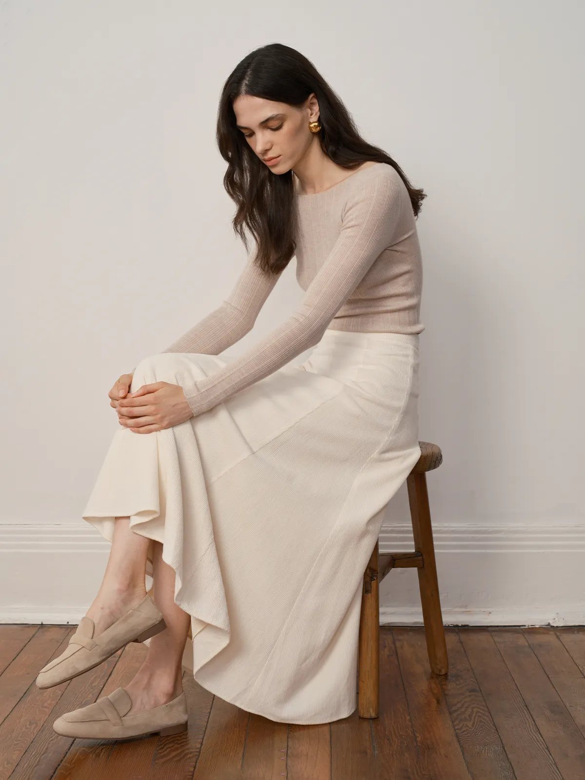 Model seated on wooden stool wearing MILIAN corduroy maxi skirt showing side drape and asymmetrical hem