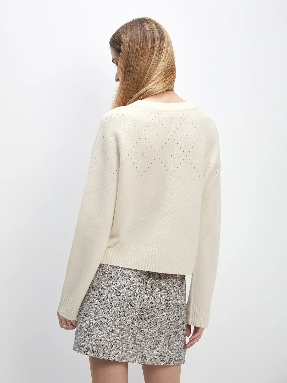 Back view of model wearing white Fair Isle hollowed knitted cardigan with diamond-shaped openwork pattern across upper back, paired with tweed mini skirt.