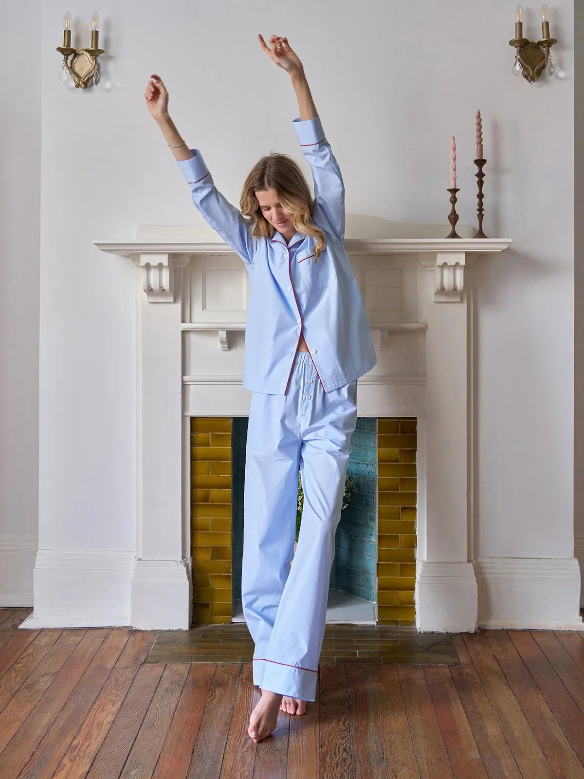 Woman stretching in light blue striped pajama set with red piping, standing before white fireplace