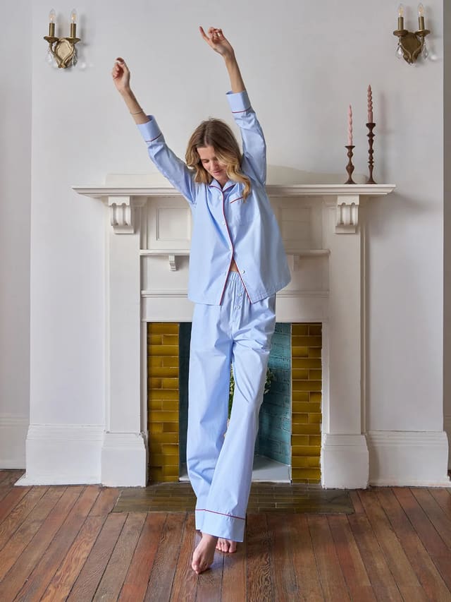 Woman stretching in light blue striped pajama set with red piping, standing before white fireplace