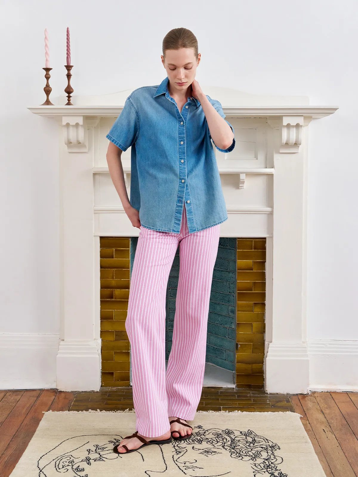 Model in chambray shirt and PINKY pink stripe jeans, standing before fireplace with candlesticks