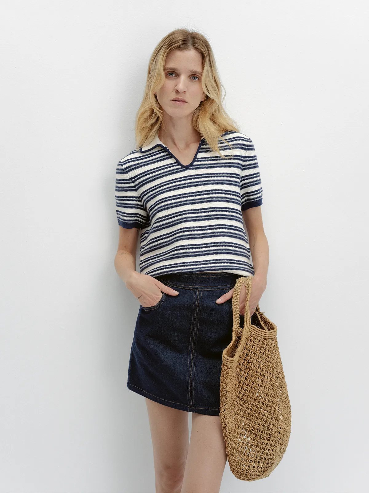 Model wearing ANGELINA striped knit polo with denim mini skirt and straw tote, hands in pockets