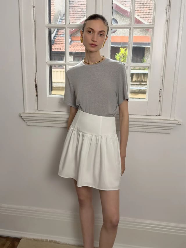 Model wearing gray MARY Lyocell Relaxed T-Shirt with white high-waisted skirt, standing by vintage window—Parisian chic minimalist styling