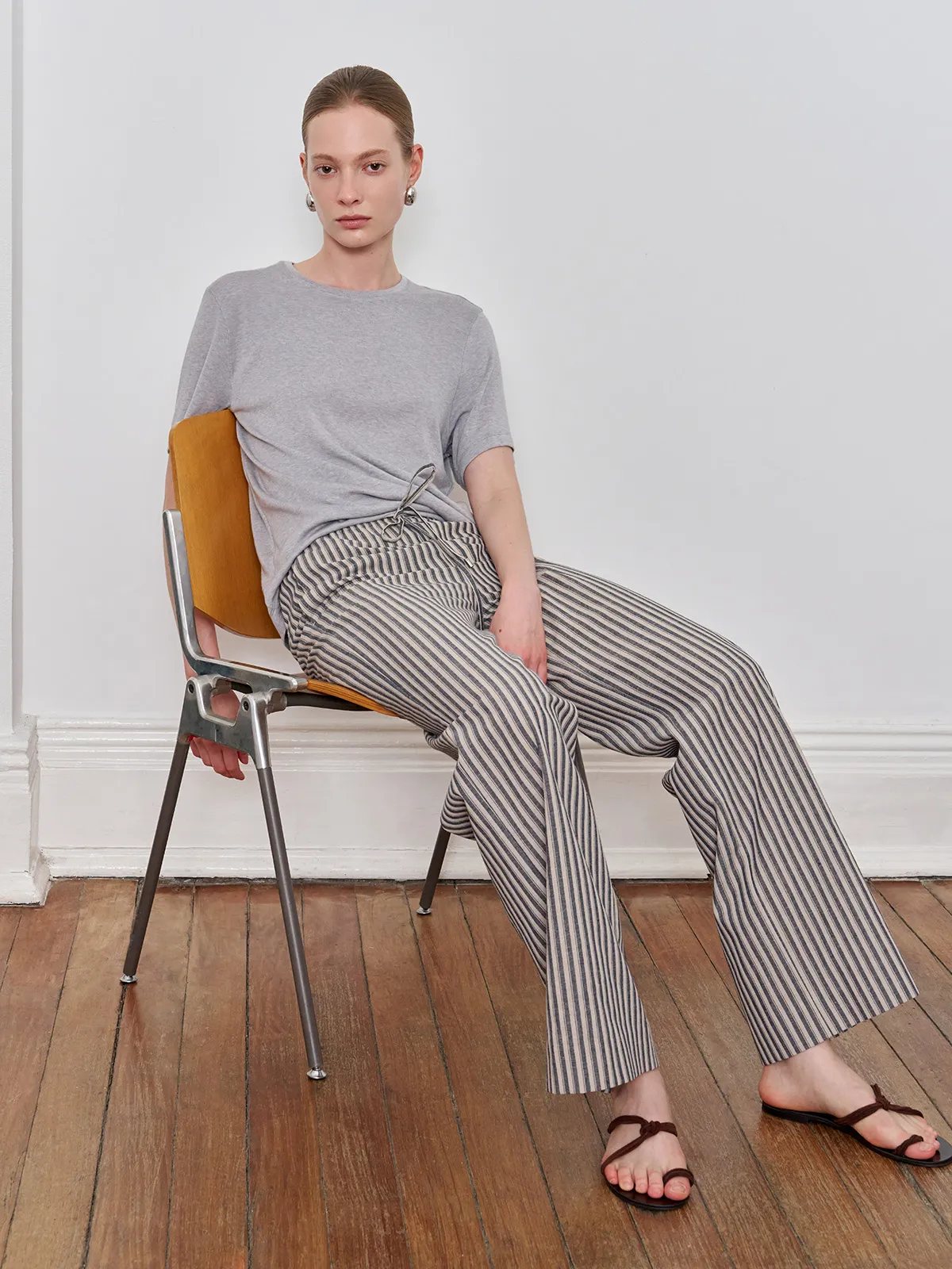 Model seated on mid-century chair wearing WHIMSY wide leg trousers with grey tee and brown sandals