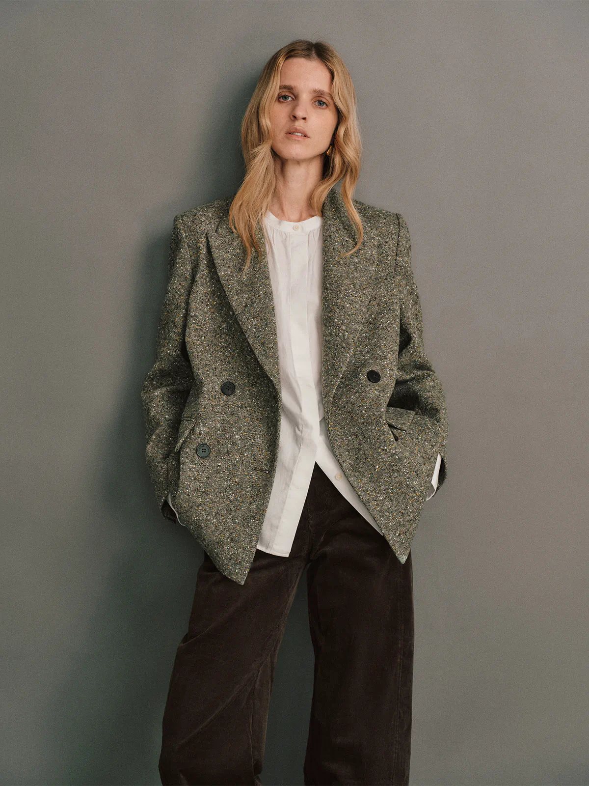 Side view of a model wearing a green wool flecked tweed double-breasted blazer, hands in pockets, with dark trousers and a top.