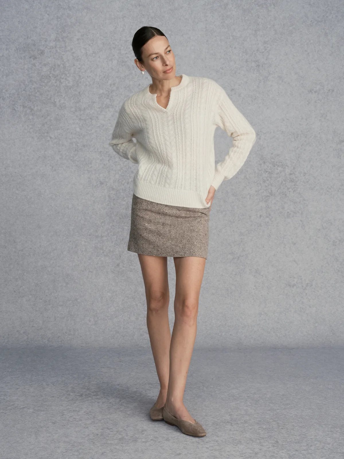 Model wearing KATIE skirt with ivory sweater, turned slightly sideways, one hand on hip, gray studio background