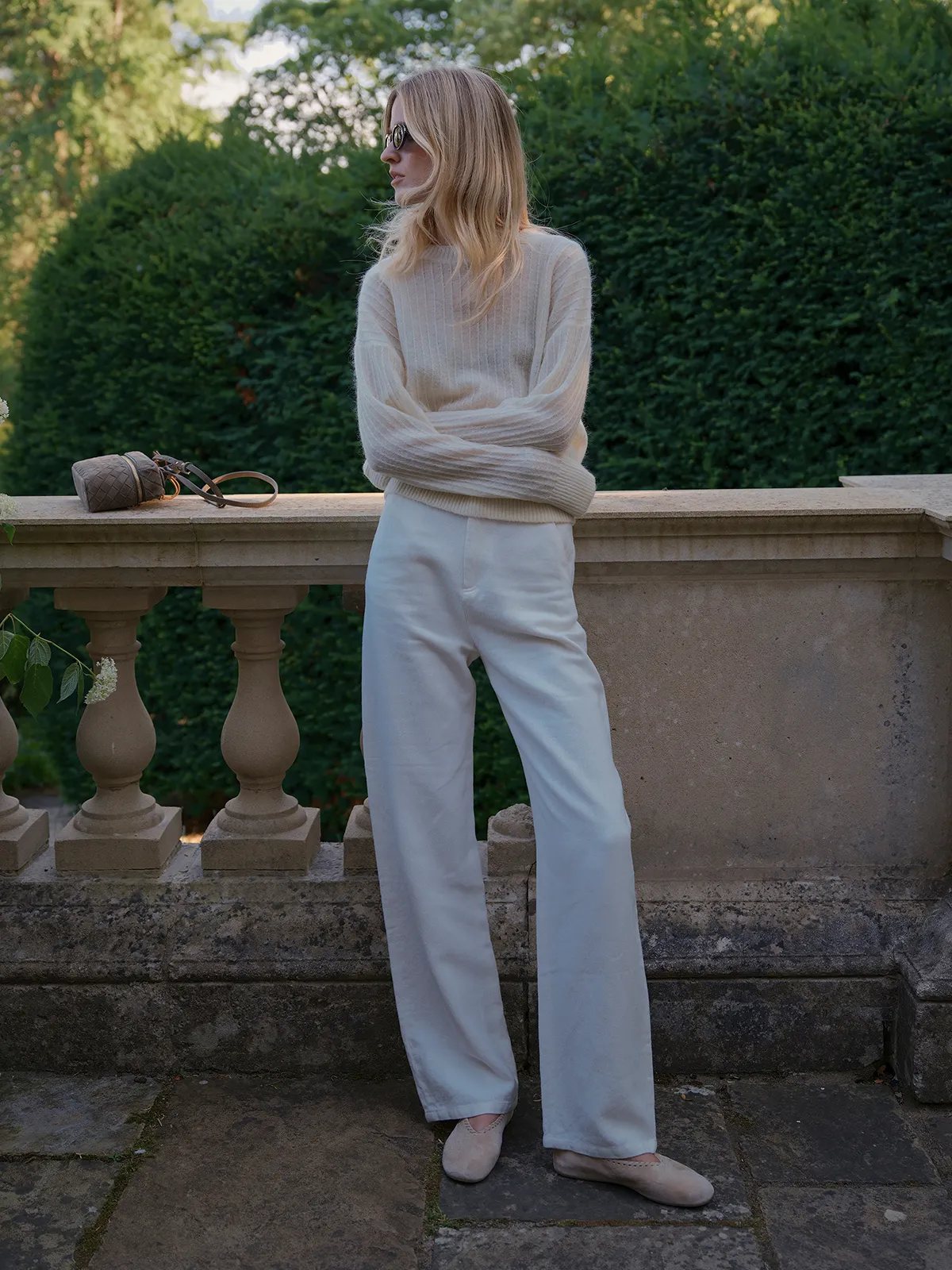 Model in ivory ELFIE jumper and white wide-leg trousers leaning on stone balustrade, sunglasses and woven bag