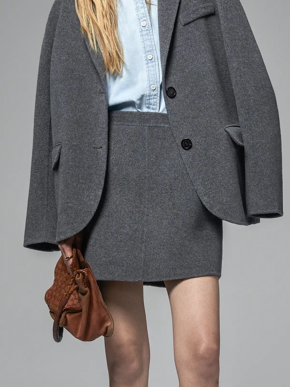 Mixed Gray short A-line skirt layered under a matching gray blazer, holding a woven bag.