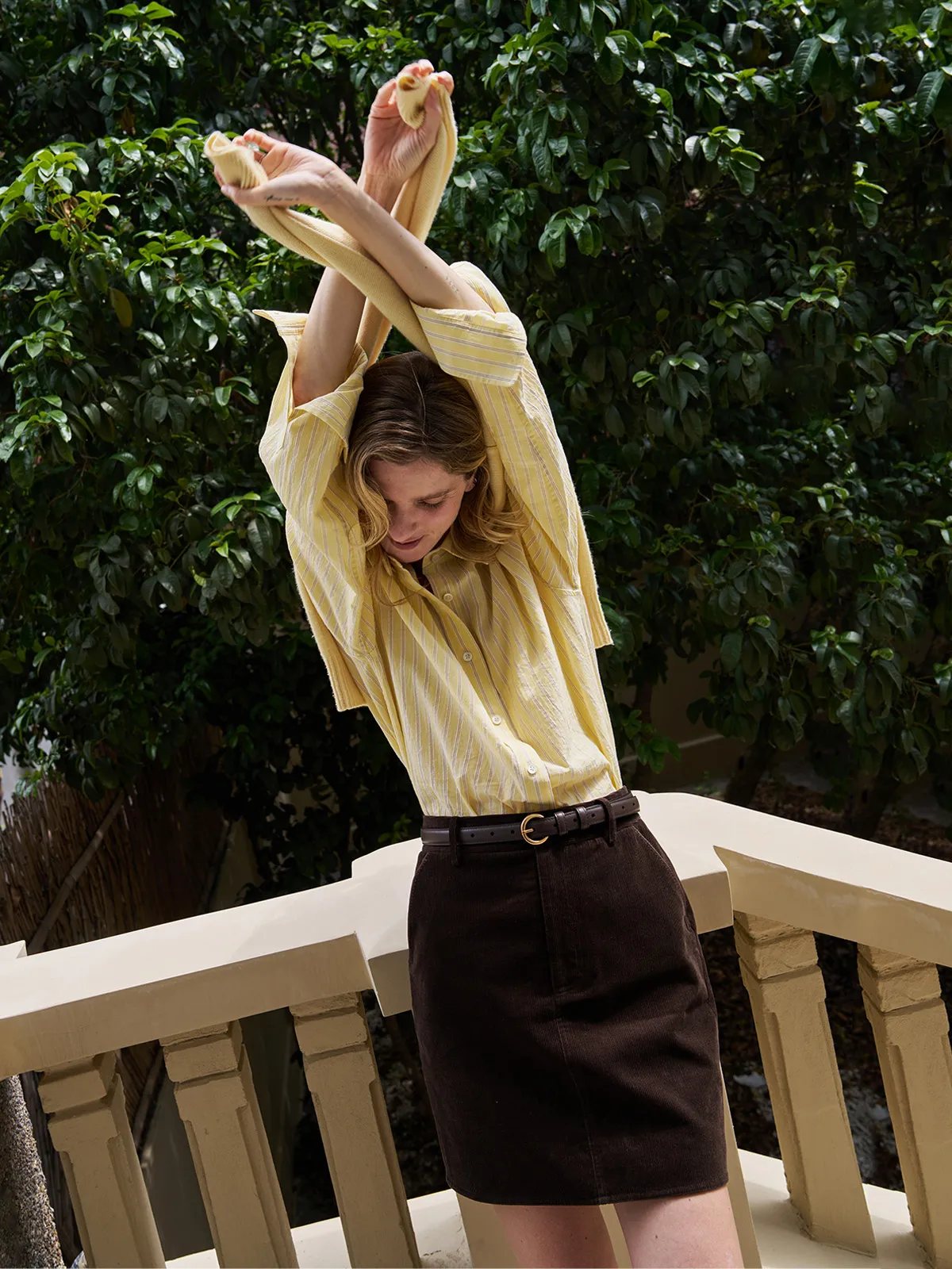 Woman wearing buttercup seersucker stripe shirt with brown corduroy skirt on balcony, quiet luxury styling
