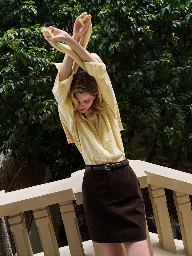 Woman wearing buttercup seersucker stripe shirt with brown corduroy skirt on balcony, quiet luxury styling
