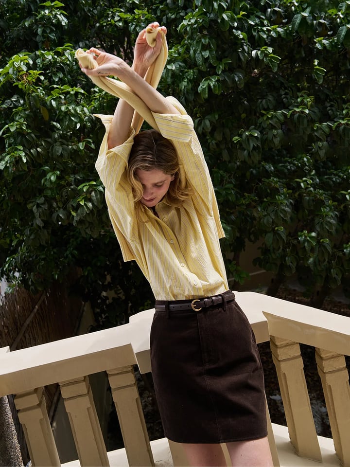 Woman wearing buttercup seersucker stripe shirt with brown corduroy skirt on balcony, quiet luxury styling