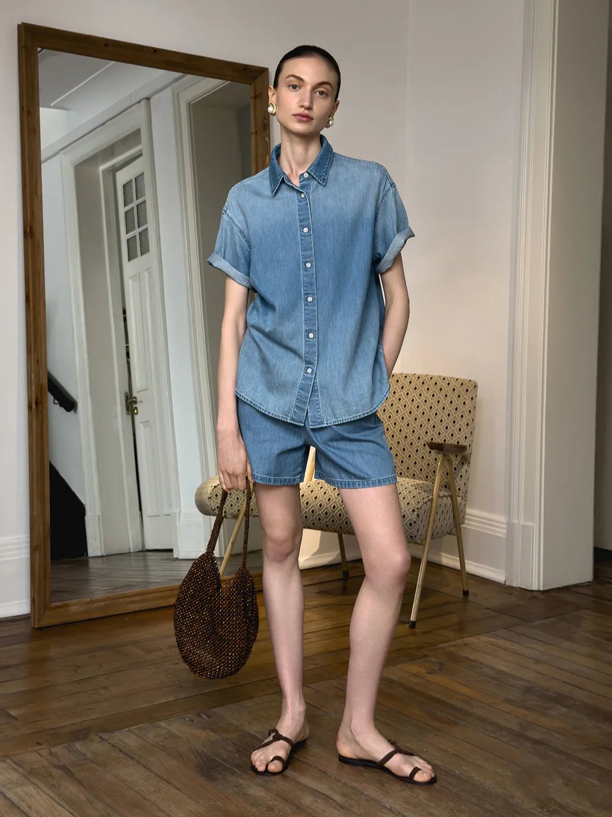 Model in denim shirt and WAYLON shorts holding beaded woven tote, standing beside floor mirror and patterned chair