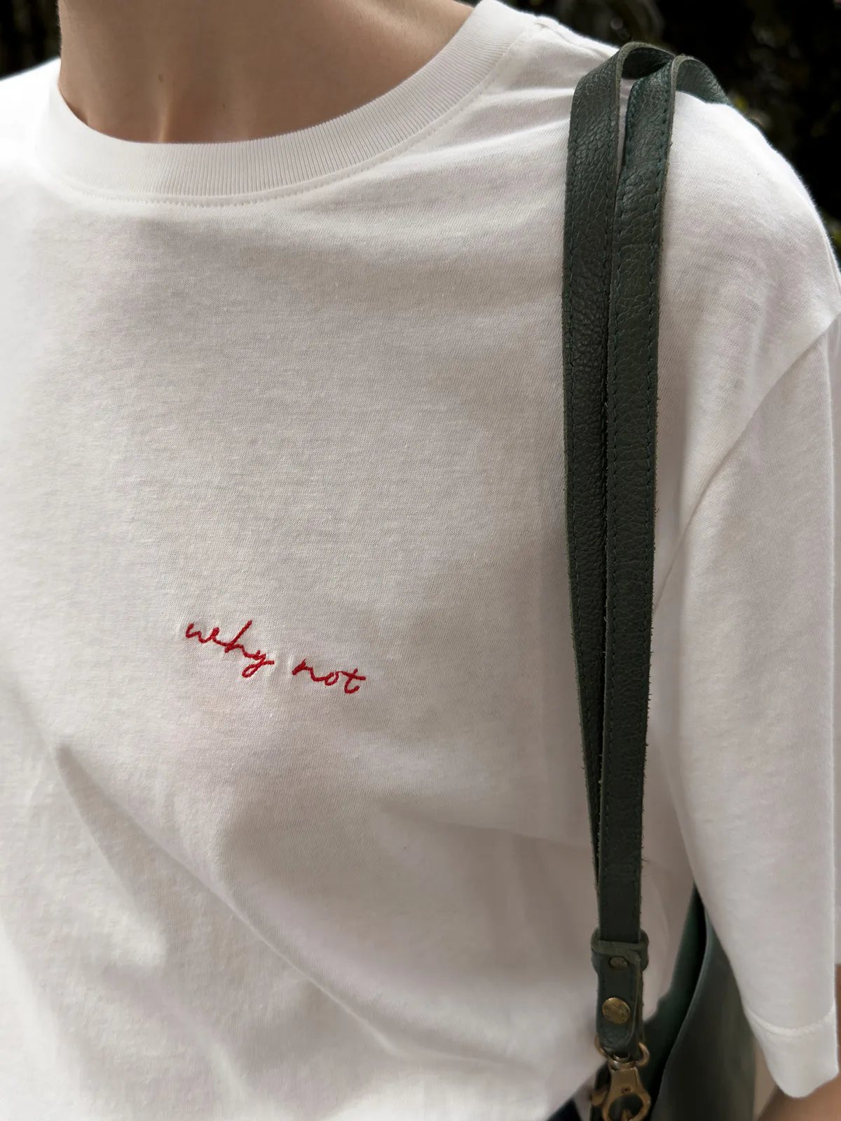 Side view of model wearing white 'why not' tee with green leather shoulder bag strap across chest