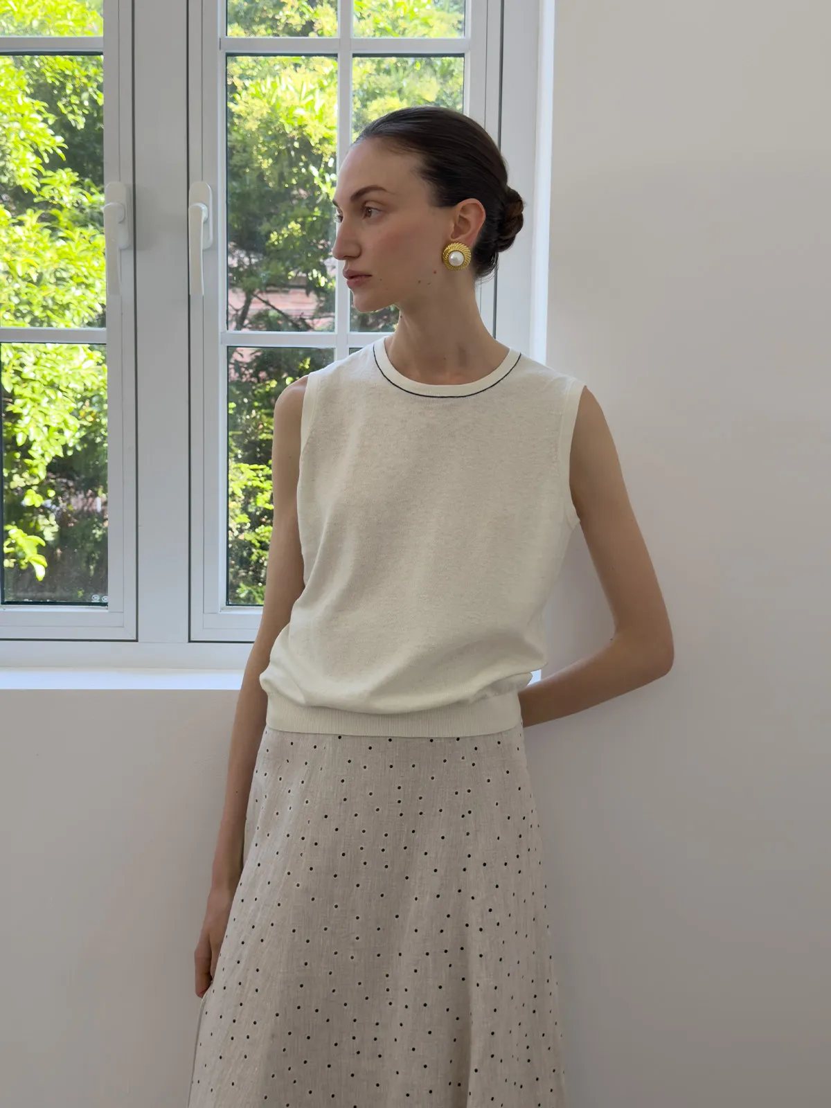 Model by window in ivory sleeveless knit top paired with polka-dot linen skirt