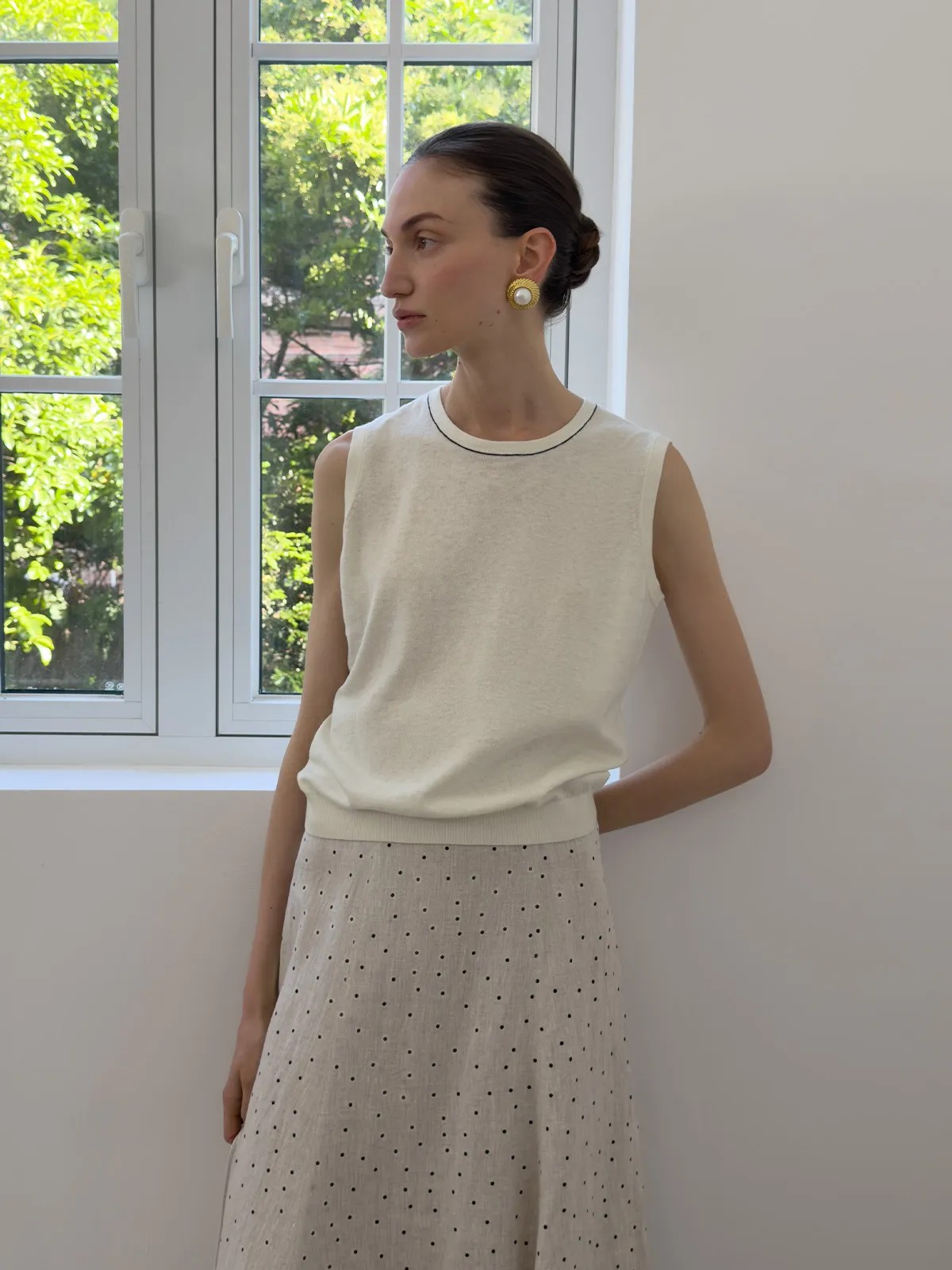 Model by window in ivory sleeveless knit top paired with polka-dot linen skirt