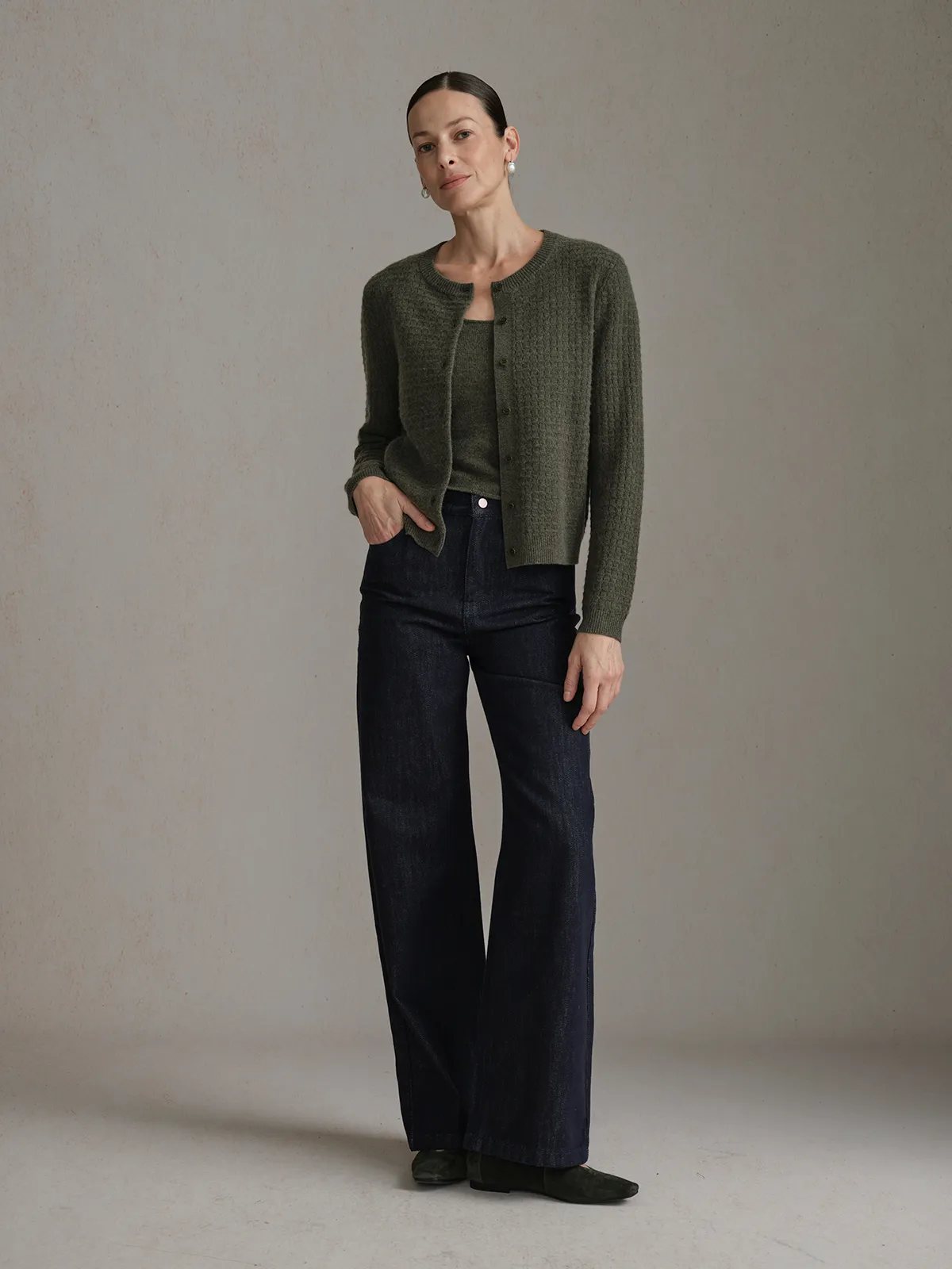 Model in CLOVER jeans with olive green textured cardigan, hands in pockets, neutral studio setting
