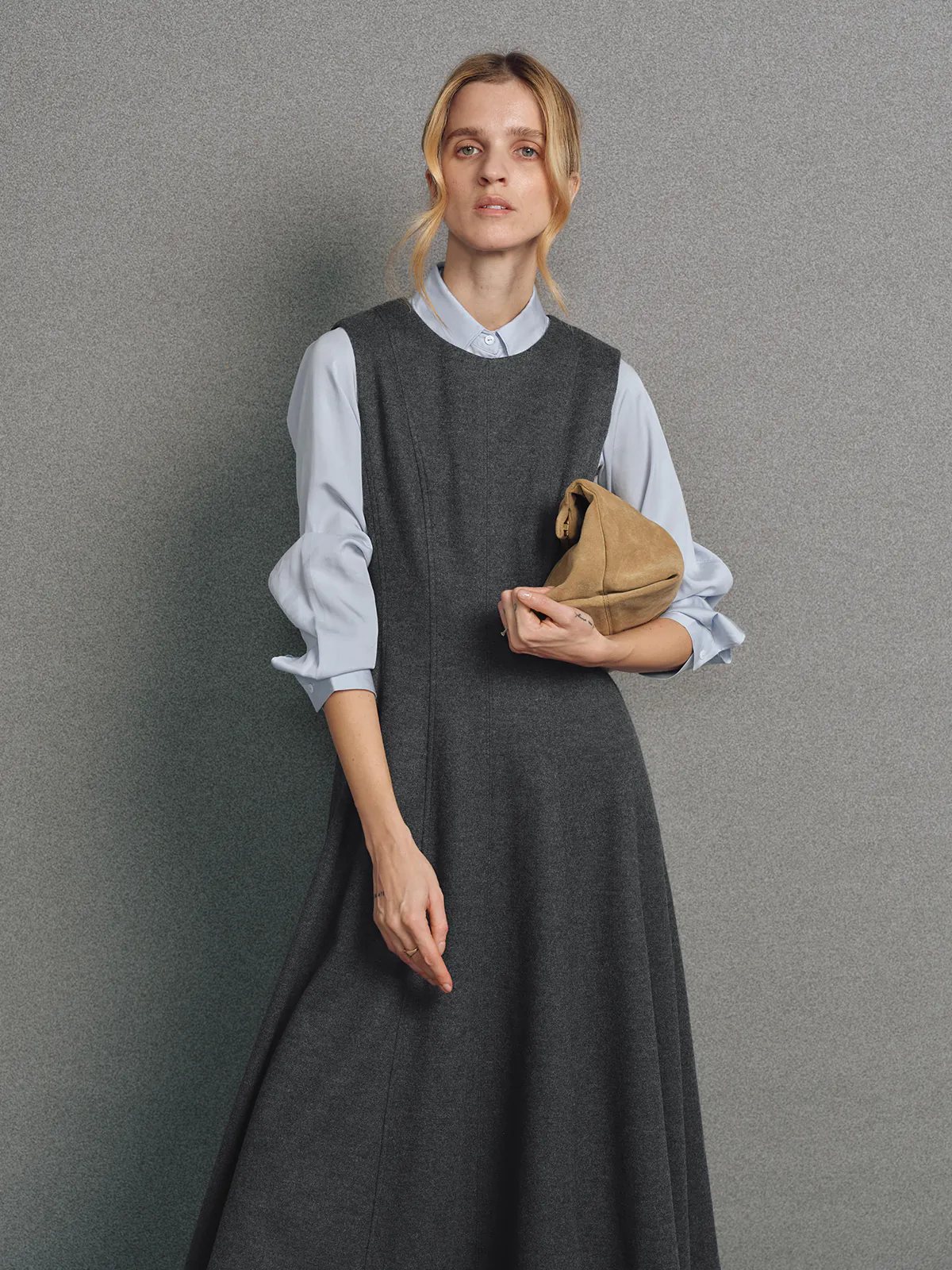 Model wearing a sleeveless dark gray wool knit A-line dress layered over a light collared shirt, holding a clutch.