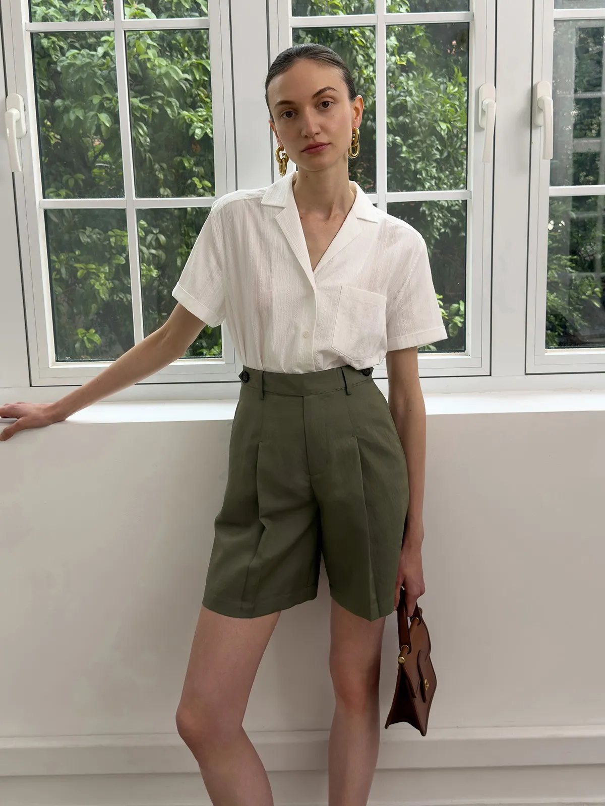 Model in olive green ADELE Bermuda shorts with white shirt, gold earrings, and brown bag by window