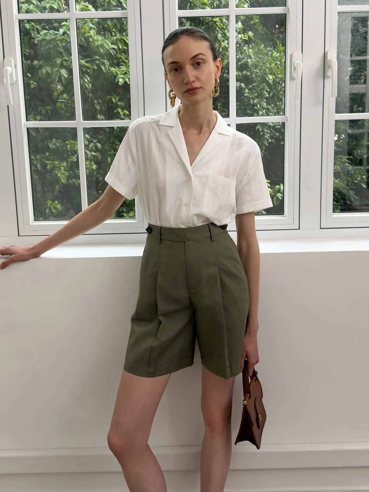 Model in olive green ADELE Bermuda shorts with white shirt, gold earrings, and brown bag by window