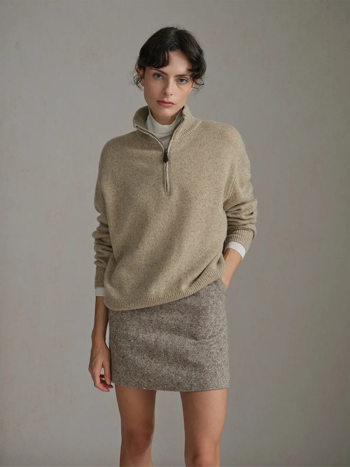 Front-facing model in KATIE skirt and beige half-zip sweater, one hand in pocket, neutral expression, soft lighting