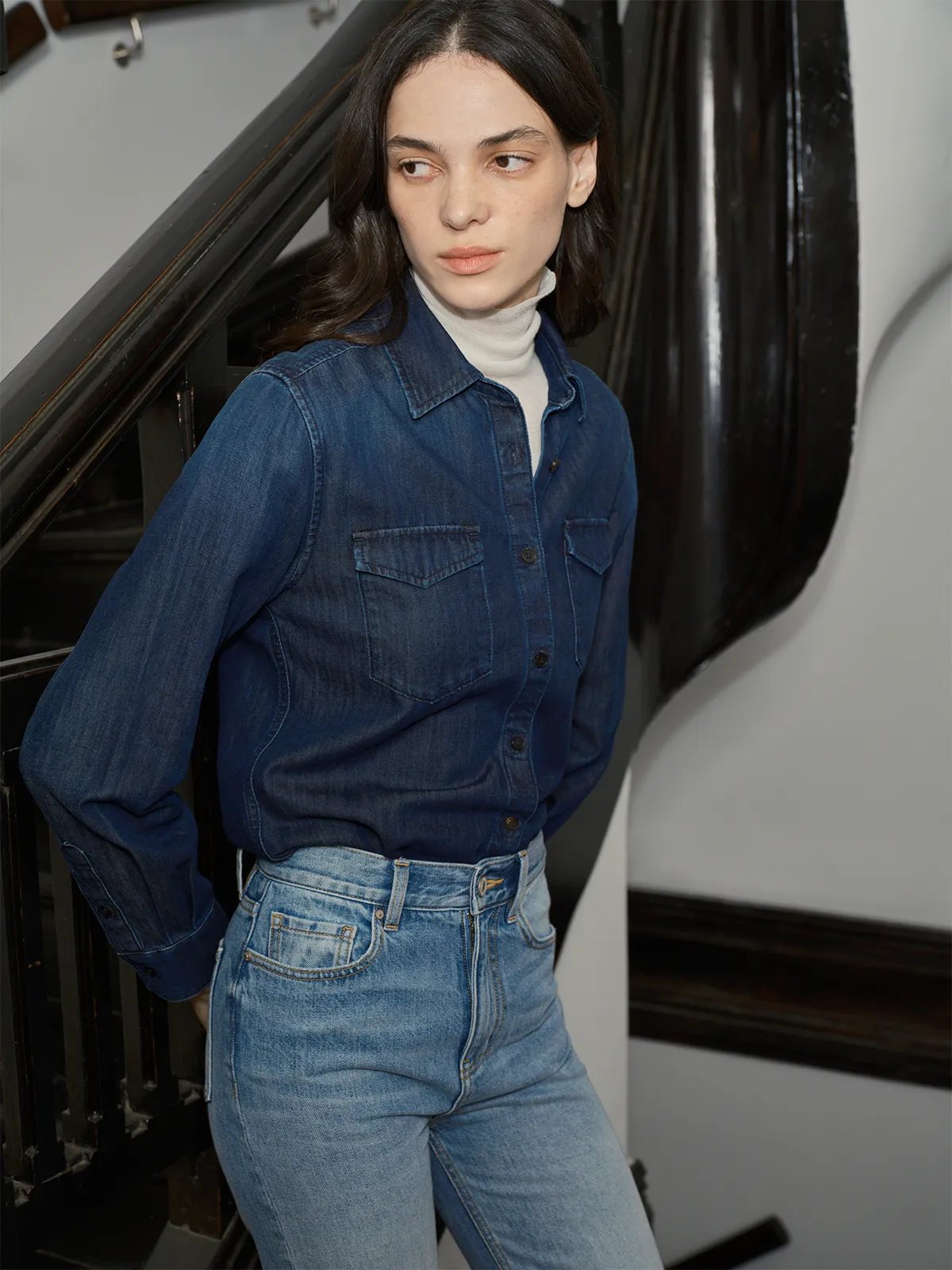 Model wearing CINDY denim shirt layered over ivory turtleneck with light wash jeans, staircase backdrop