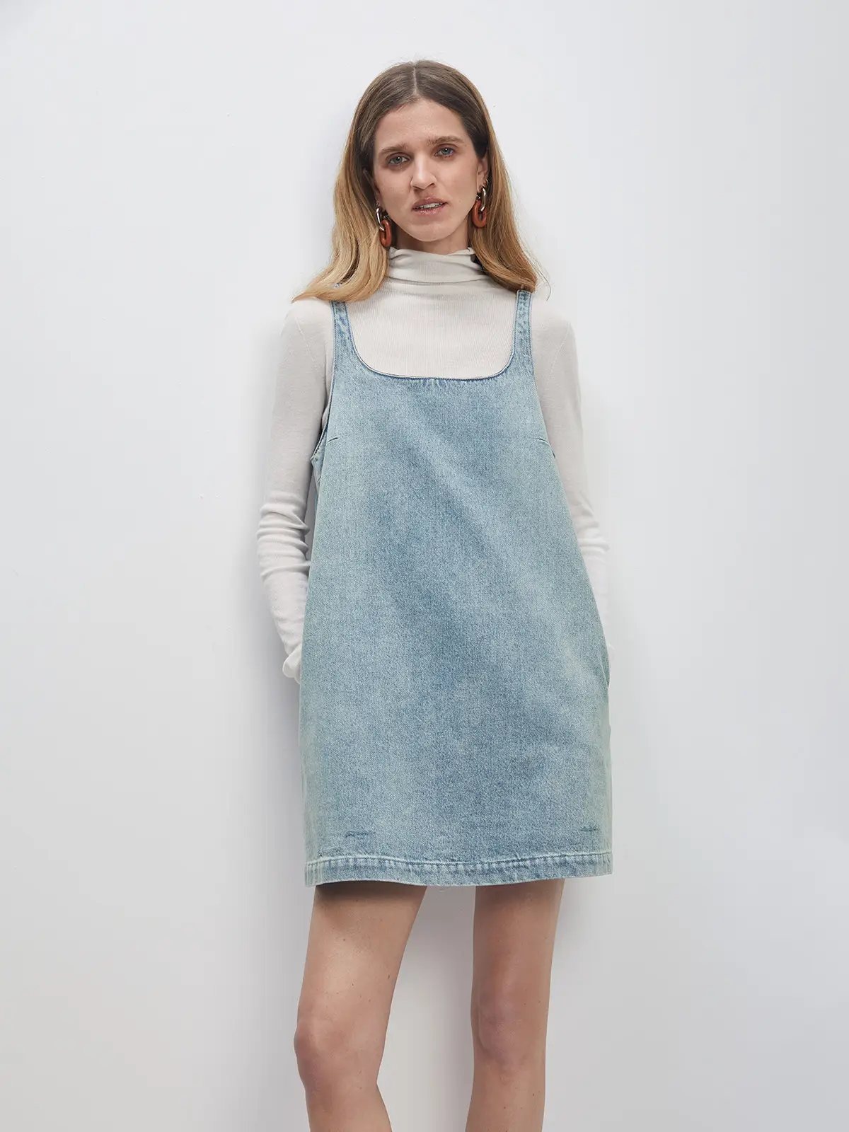 Model wearing a Sky Blue A-line denim dress over a turtleneck, hands in pockets.