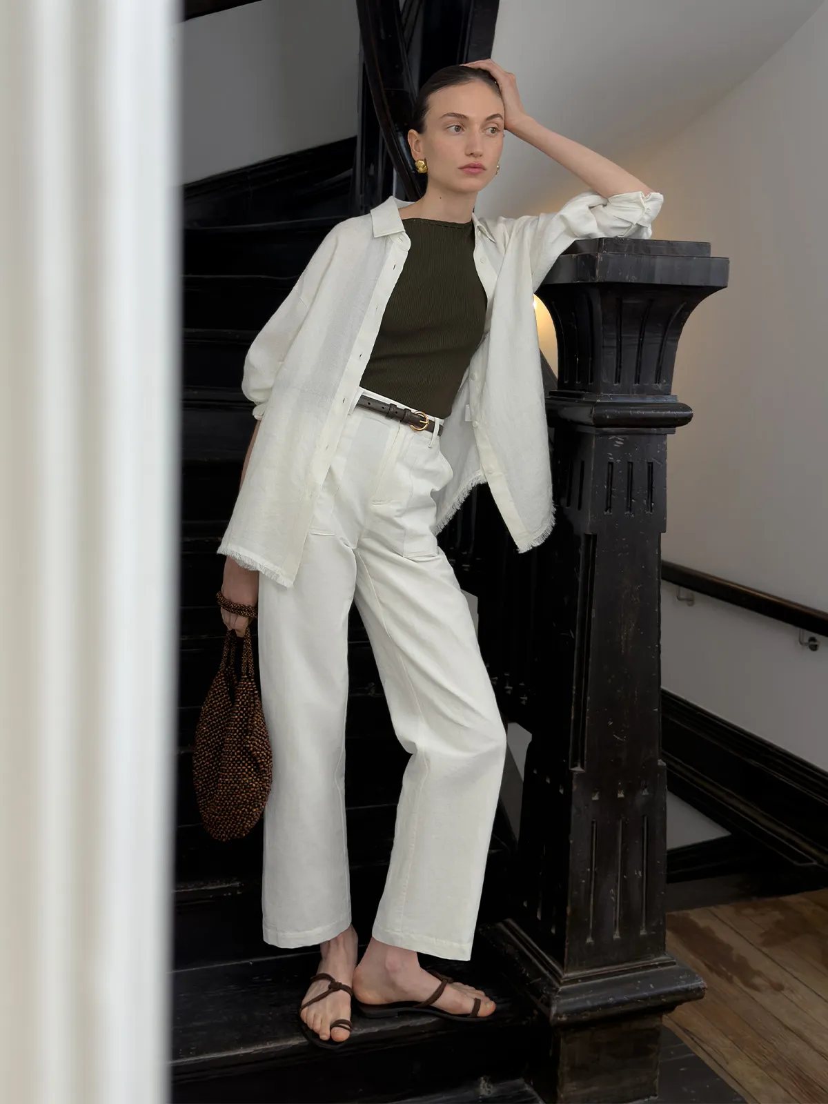 Model wearing ELARA shirt open over olive ribbed tank, belted white trousers, on staircase