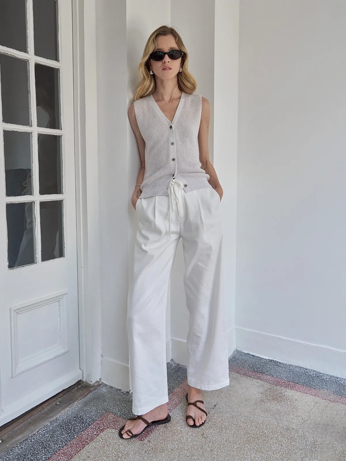Model wearing white LEIGHTON linen-cotton curved leg trousers with sleeveless knit vest and sandals against white doorframe
