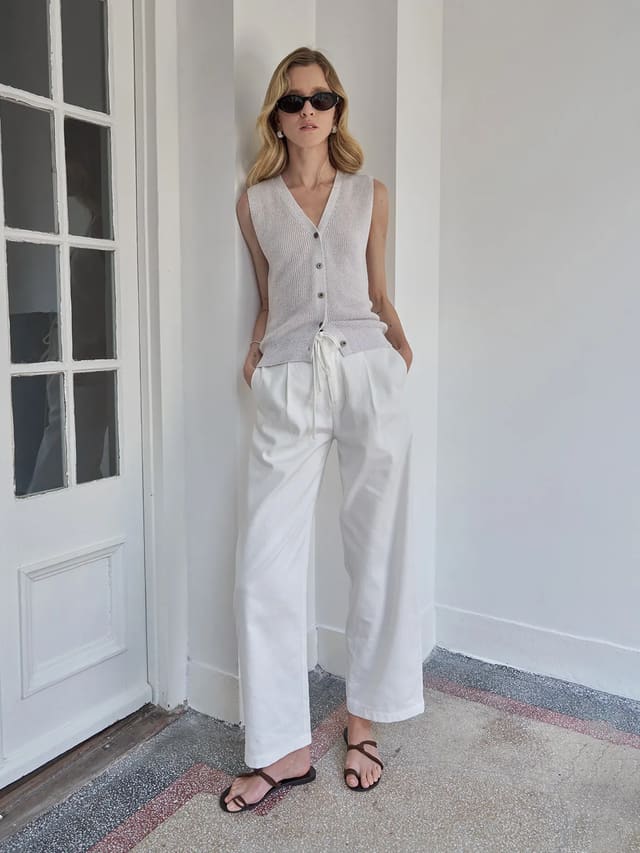 Model wearing white LEIGHTON linen-cotton curved leg trousers with sleeveless knit vest and sandals against white doorframe