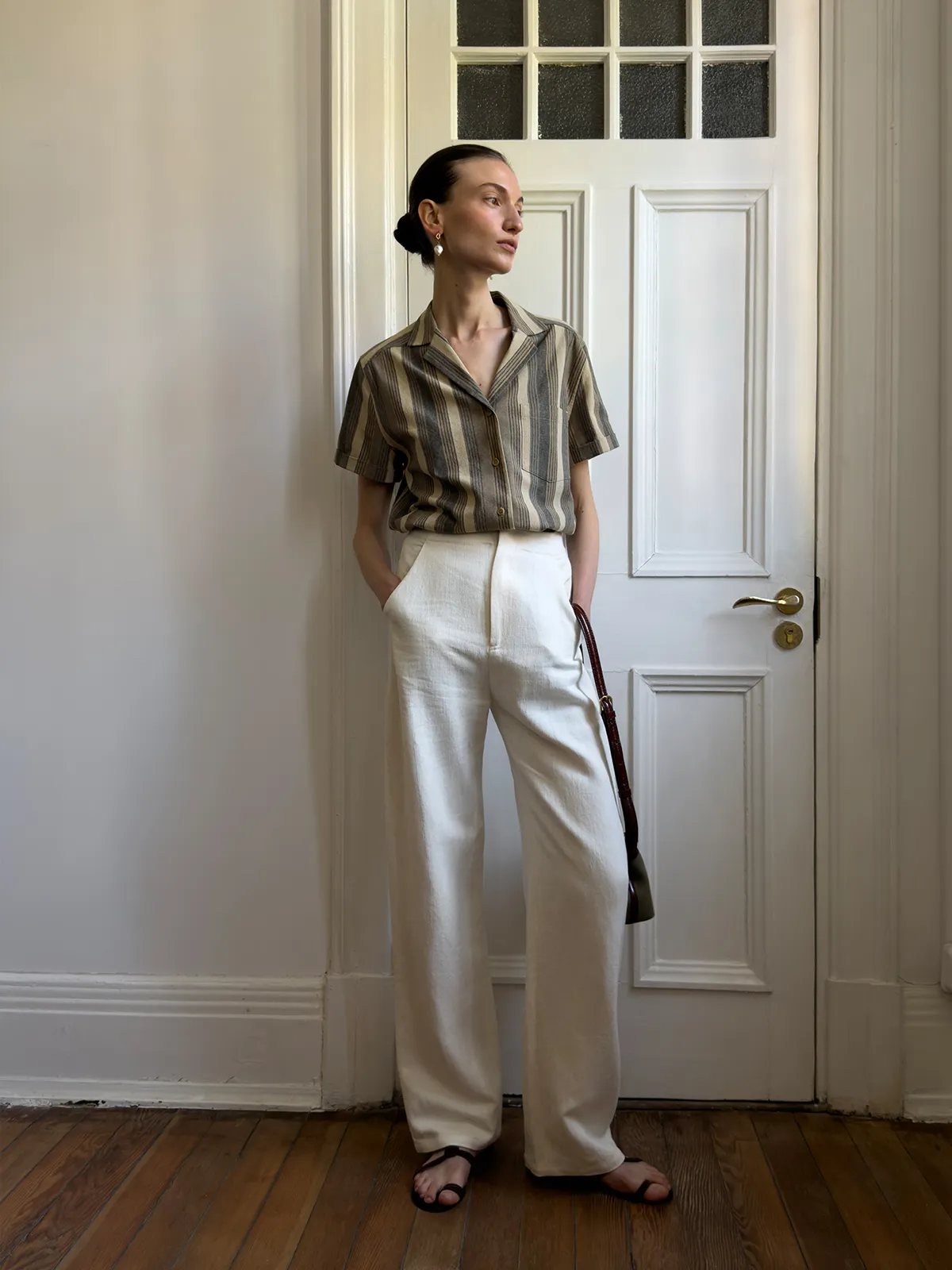 Model wearing REAGAN shirt with cream wide-leg trousers and brown leather bag by white door
