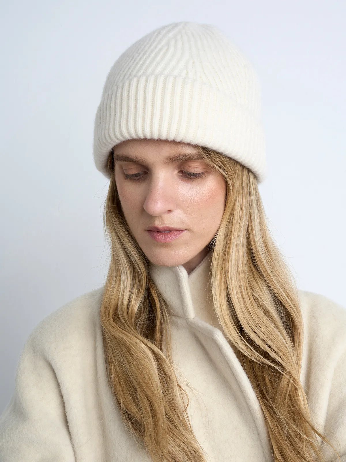 Close-up front view of a white ribbed knit beanie worn by a model with long blonde hair and coat.
