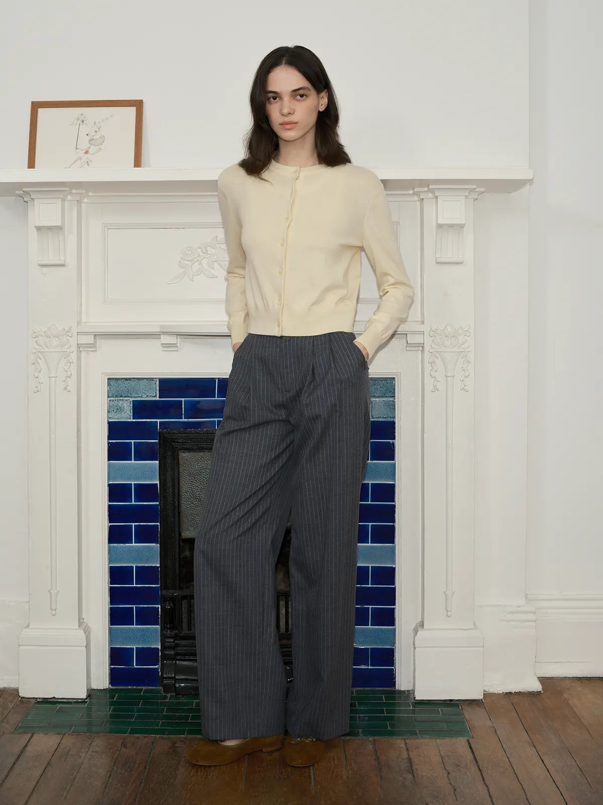 Front-facing model in BINX cardigan with hands in pockets, standing before blue-tiled fireplace