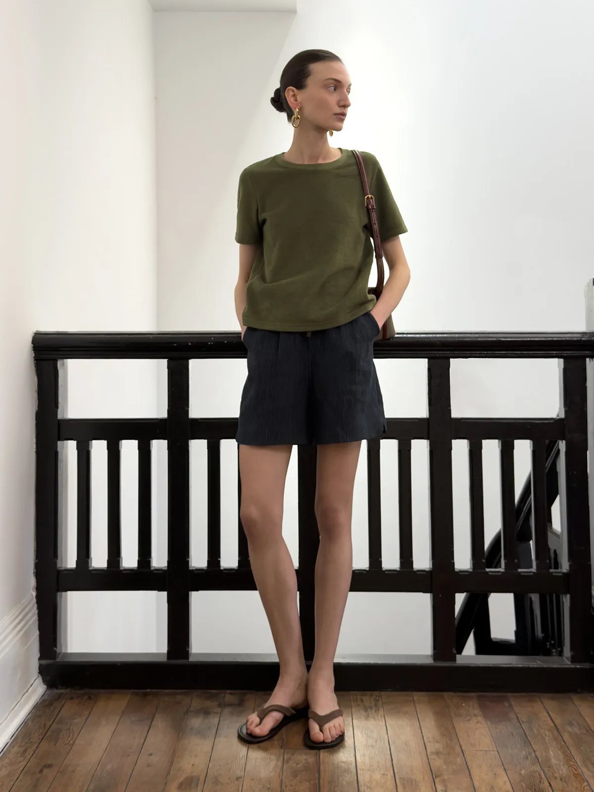Model in olive green tee and NEVAEH shorts leaning against black railing, wearing brown sandals