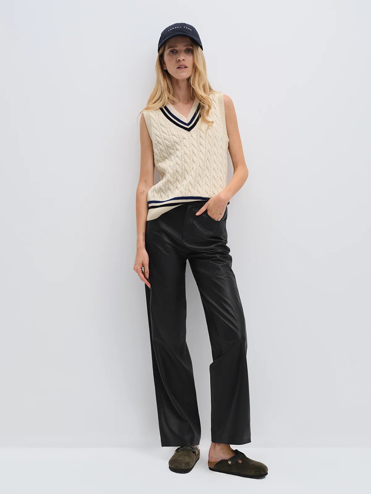 Model wearing andreatang wool-cotton cable-knit cricket vest with contrast stripes, styled with black leather pants and cap
