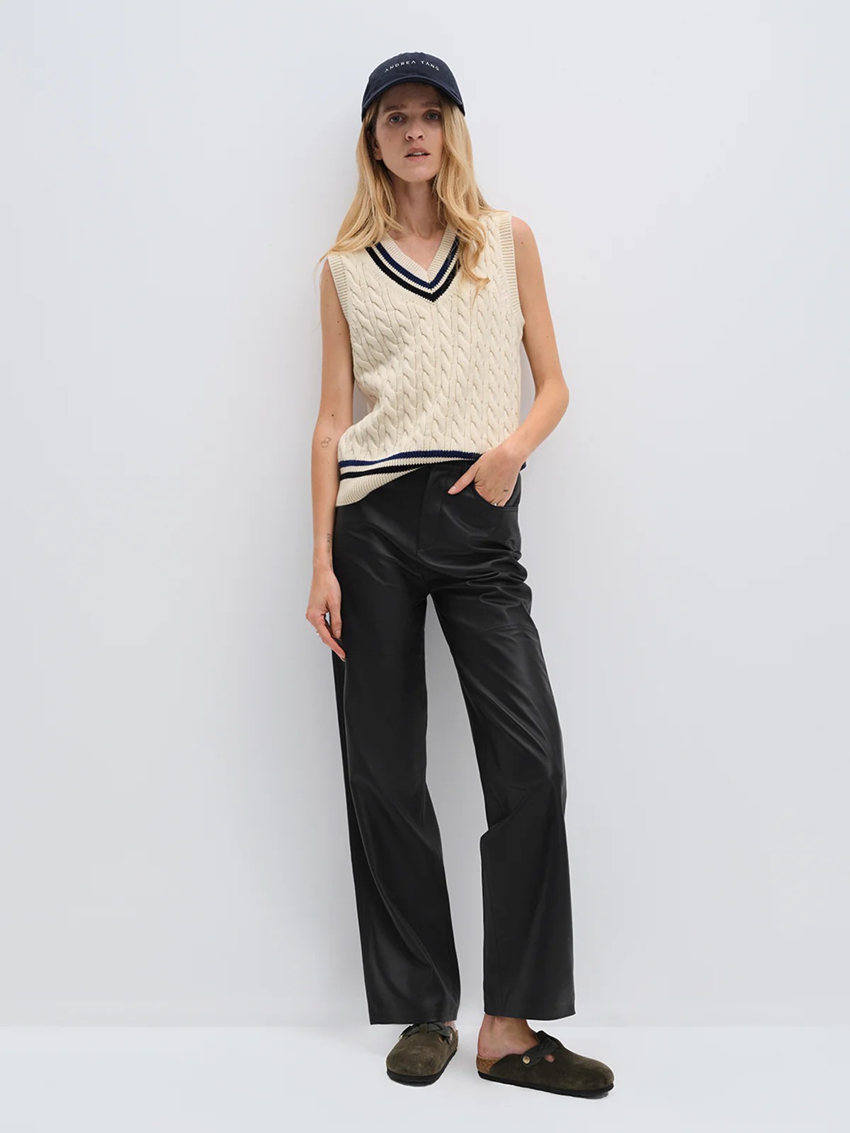 Model wearing andreatang wool-cotton cable-knit cricket vest with contrast stripes, styled with black leather pants and cap