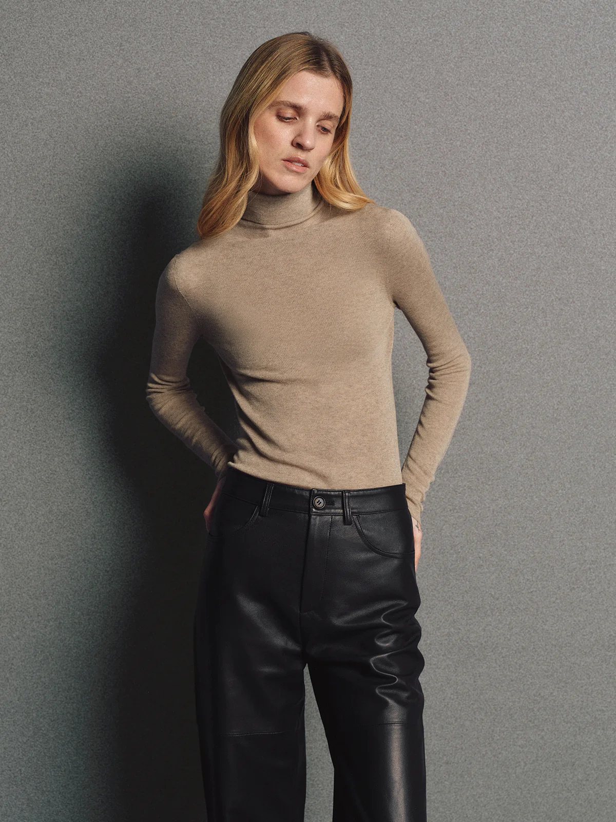 Side view of model in a seamless turtleneck in Brown, hands in pockets, with leather pants.