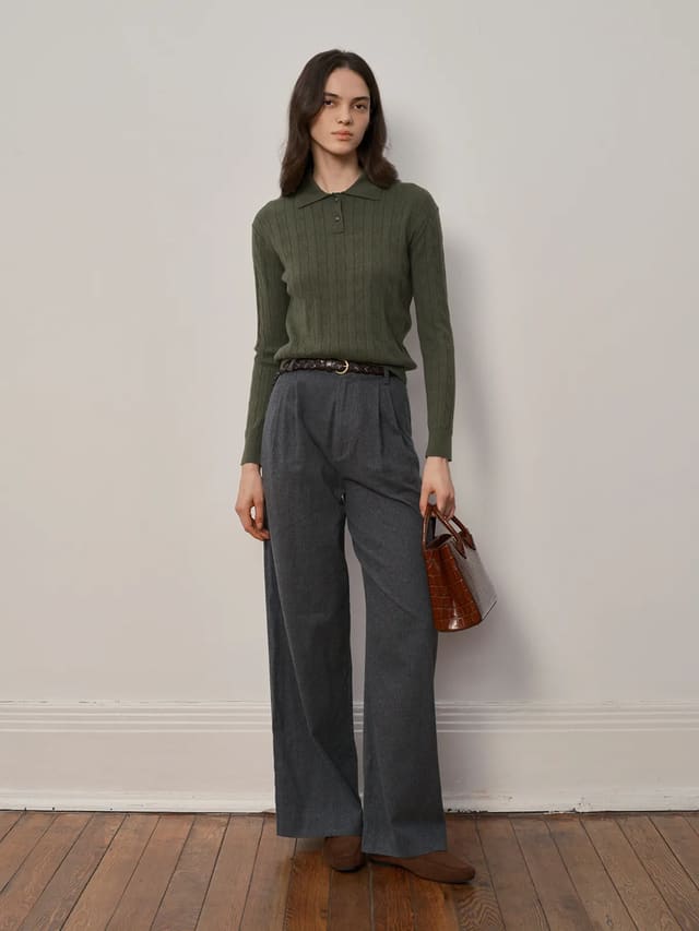 Model wearing olive green EMERSON Cable Knit Polo Jumper with grey wide-leg trousers and brown leather belt, holding croc-embossed mini bag—quiet luxury styling on wooden floor against white wall